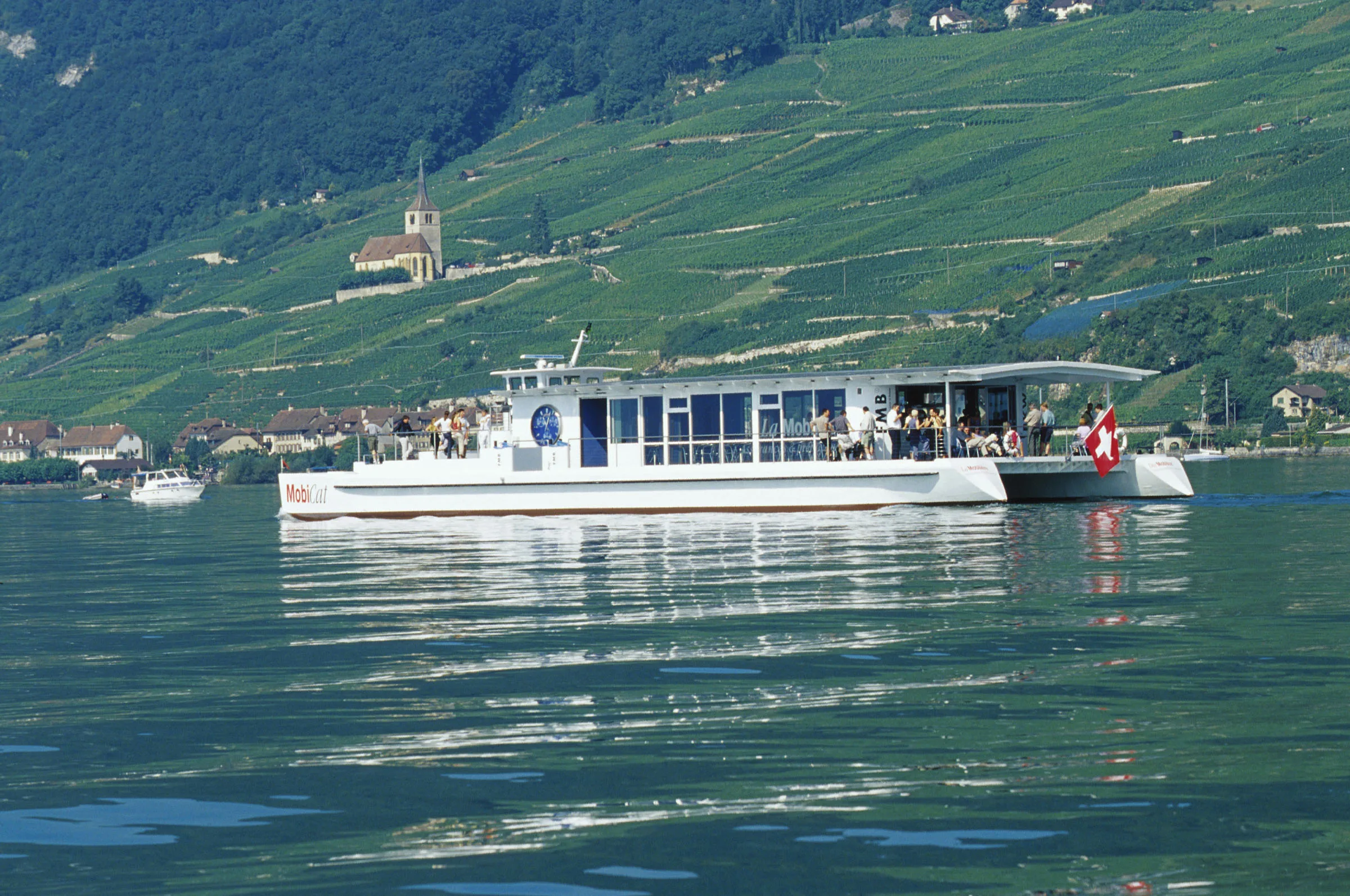The white MobiCat solar catamaran cruising on Lake Biel below vineyards near Ligerz