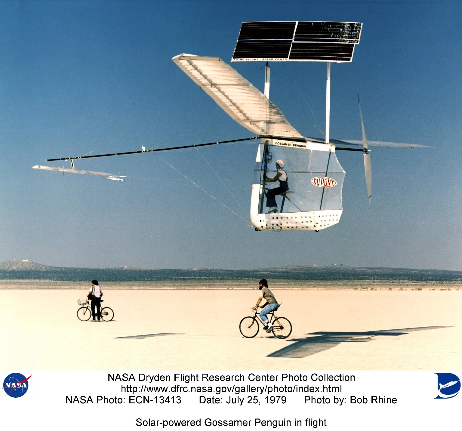 The ultralight Gossamer Penguin solar aircraft flying low over a dry lakebed with cyclists below
