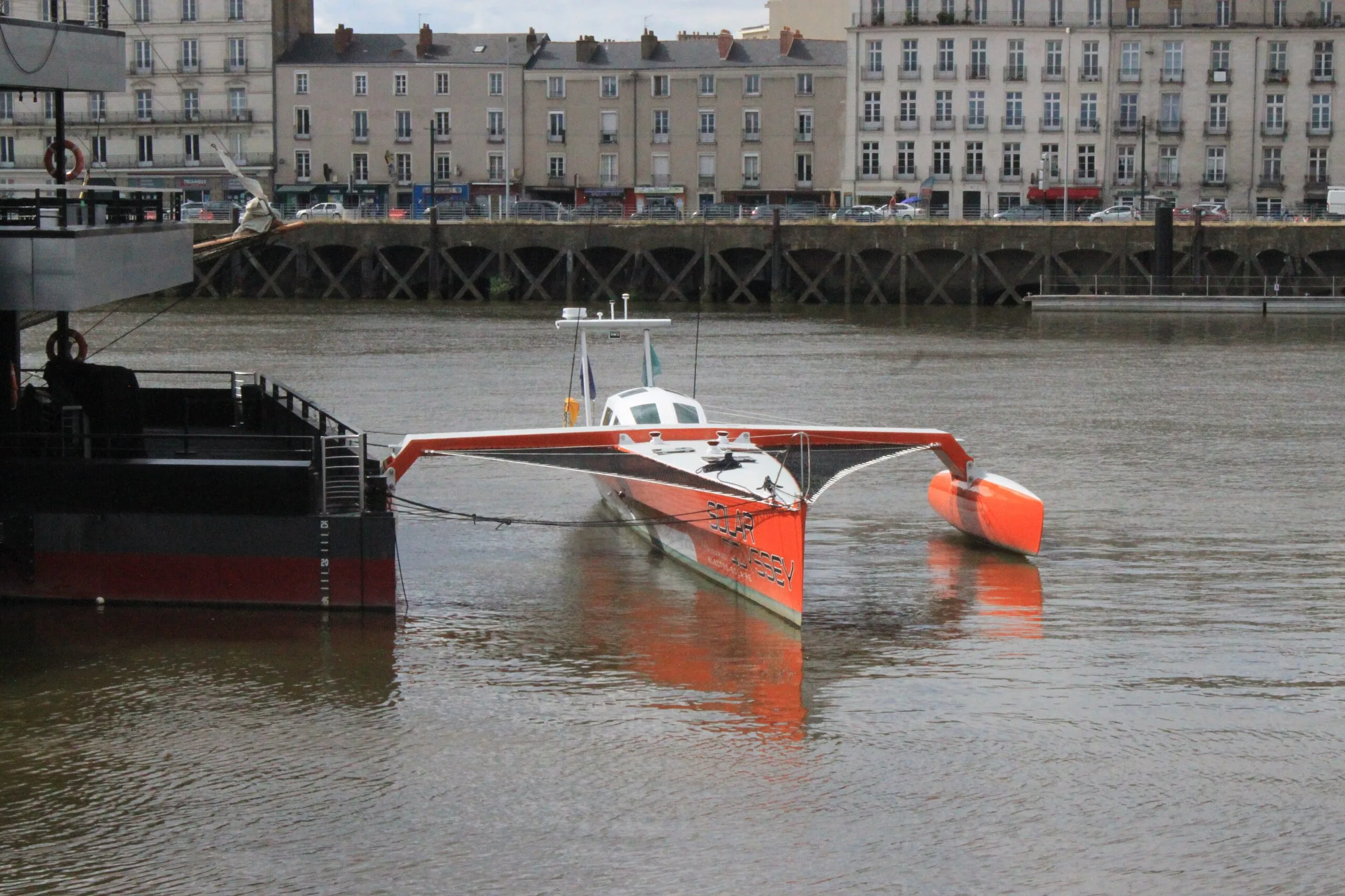 The orange and white Solar Odyssey trimaran moored on the river in Nantes